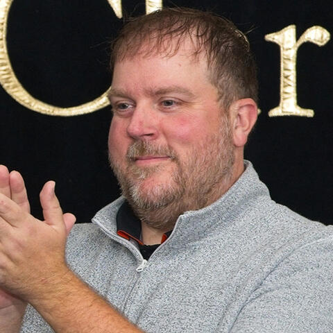 Ryan G Photo of Ryan G clapping in a grey pullover looking to the right of frame with a black background.